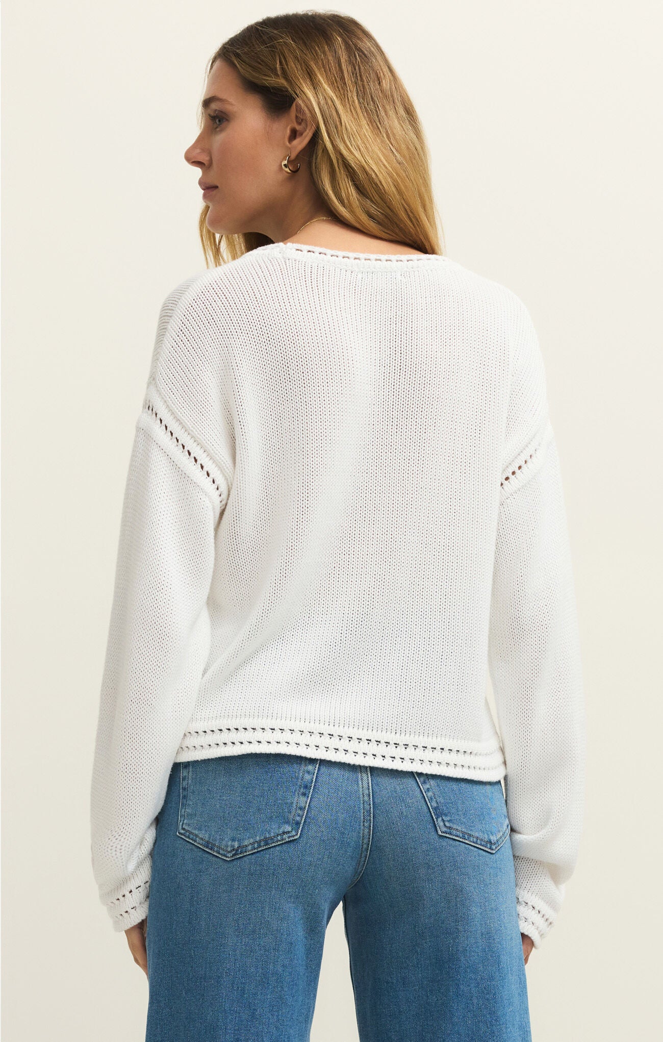 Back view of woman wearing a white sweater with lace details and blue jeans on a plain background