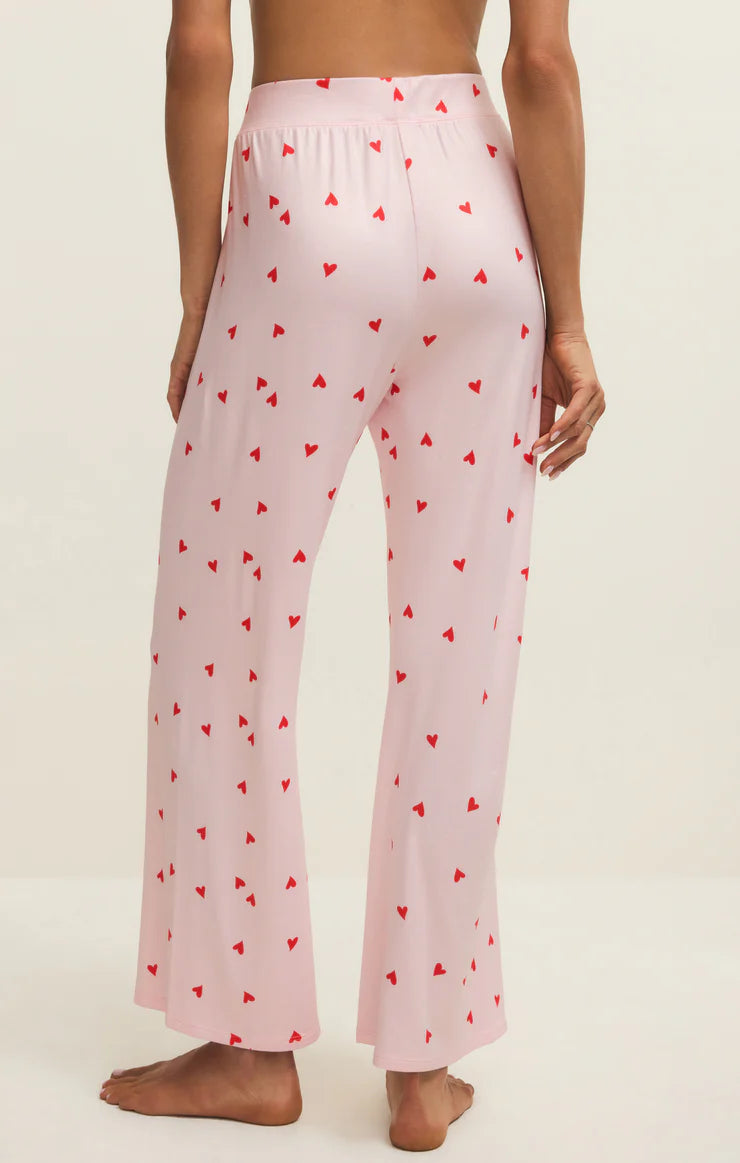 Back view of a woman wearing pink pajama pants with red heart pattern worn by a person on a white background